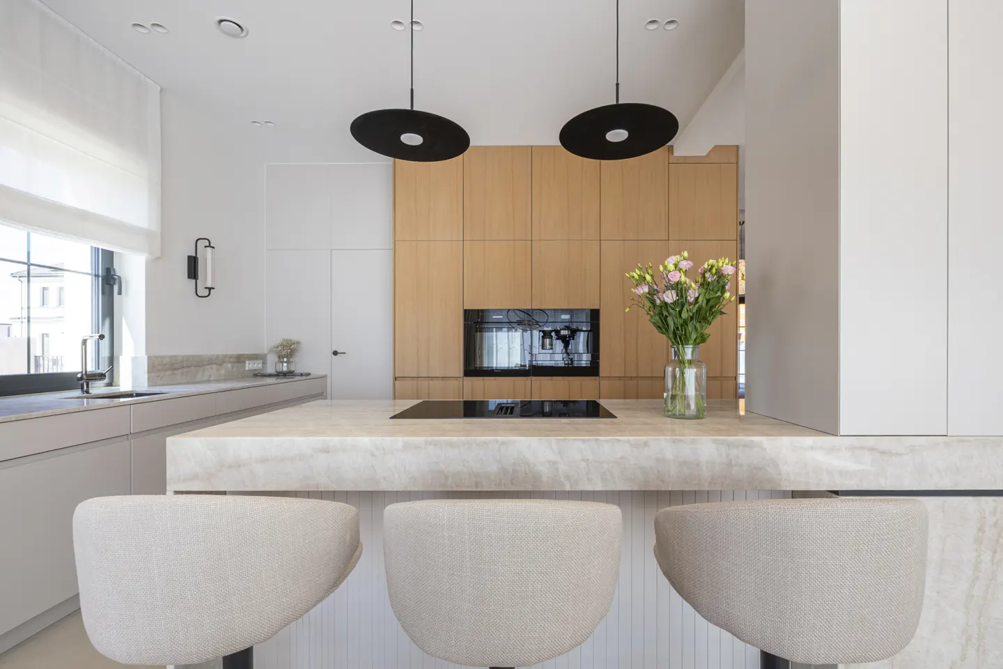 A modern kitchen with a marble island, three beige stools, and wooden cabinets. Black pendant lights hang above the island. A vase of pink flowers adds a pop of color.