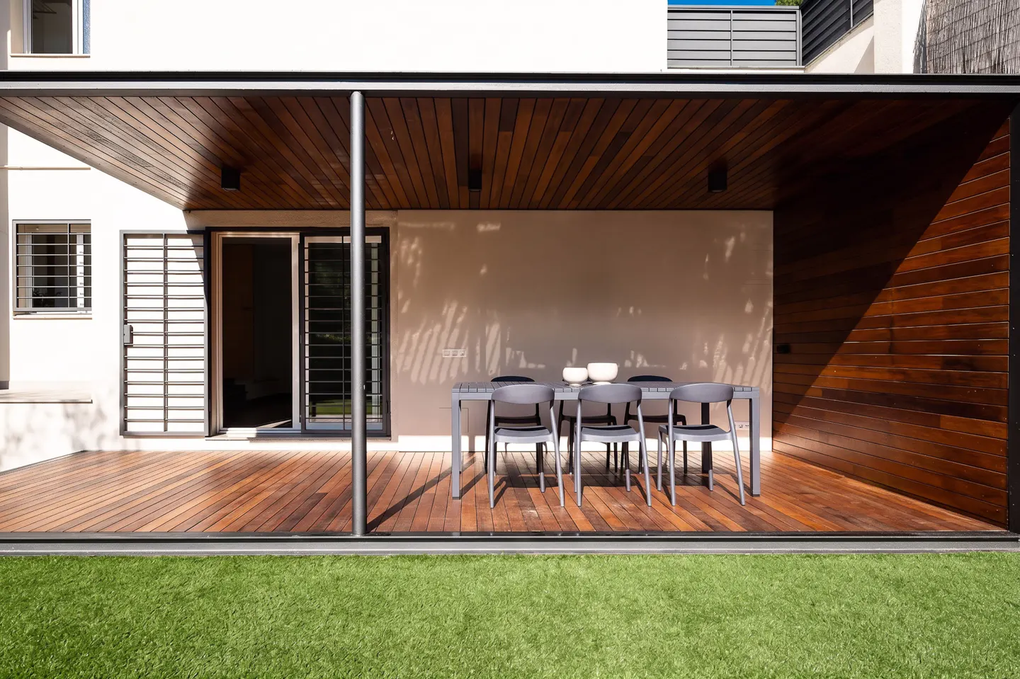 Modern patio with wooden decking, a dining set, and a covered roof, overlooking a green lawn.