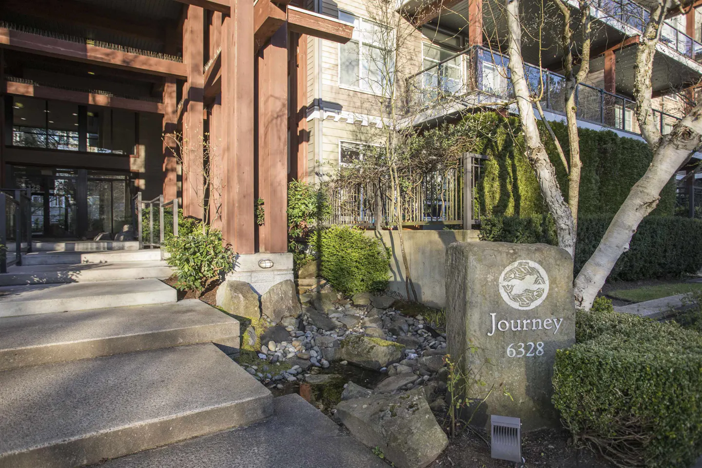 Exterior of "Journey 6328" building with stone sign, water feature, and brown wood beams.