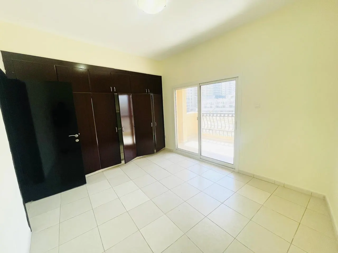 A bright, empty bedroom with cream walls, white tile floor, and dark wood built-in wardrobe. Sliding glass doors lead to a balcony.