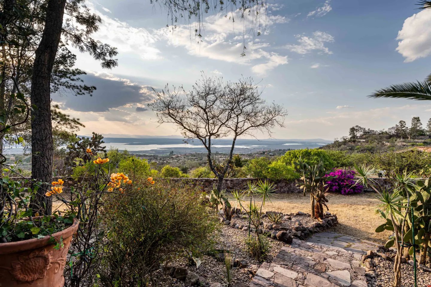Scenic view of a garden with stone path, flowers, and a distant lake under a cloudy sky.
