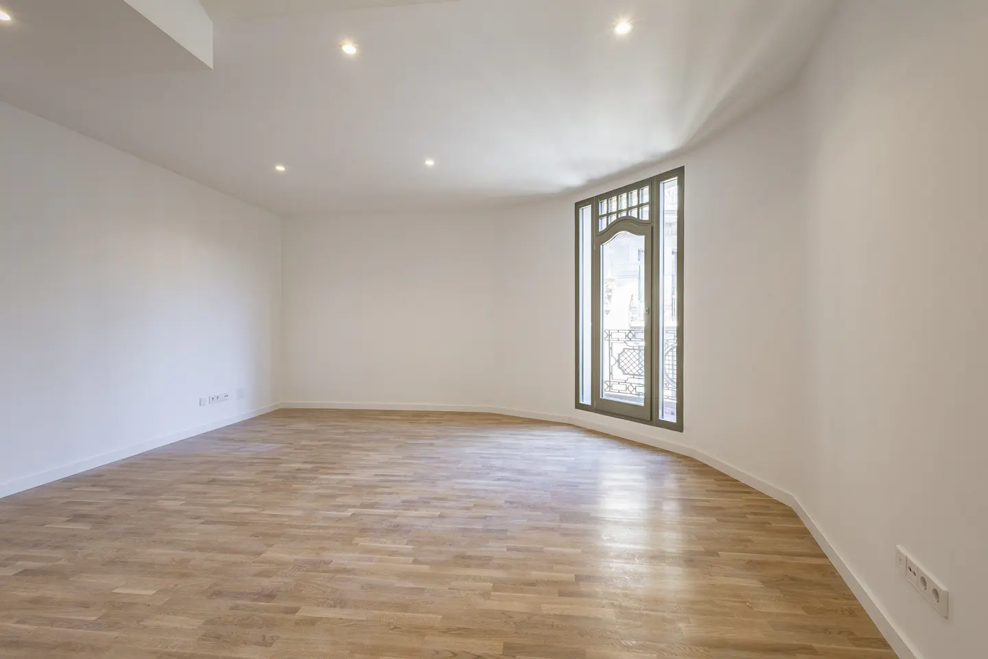 Empty room with light wood floors, white walls, and a large window with a balcony view. Recessed lighting in the ceiling.