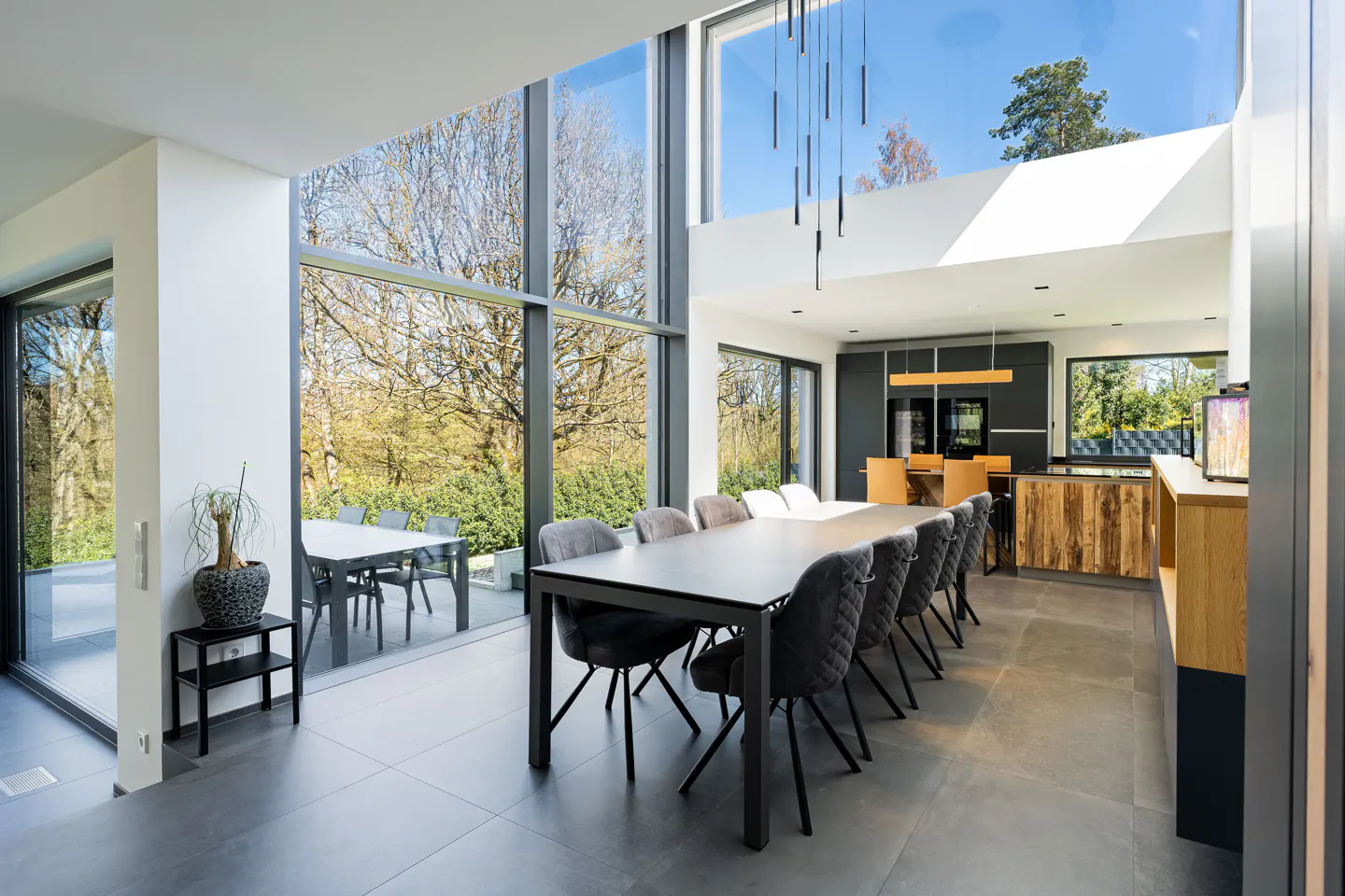 Bright, modern dining room with a long table and gray chairs. Large windows offer views of trees. Kitchen in background.