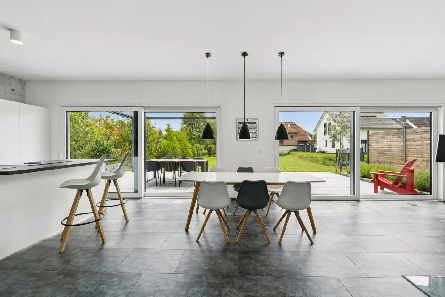 Bright, modern dining area with a white table, gray chairs, and black pendant lights. Sliding glass doors open to a green backyard with patio furniture.