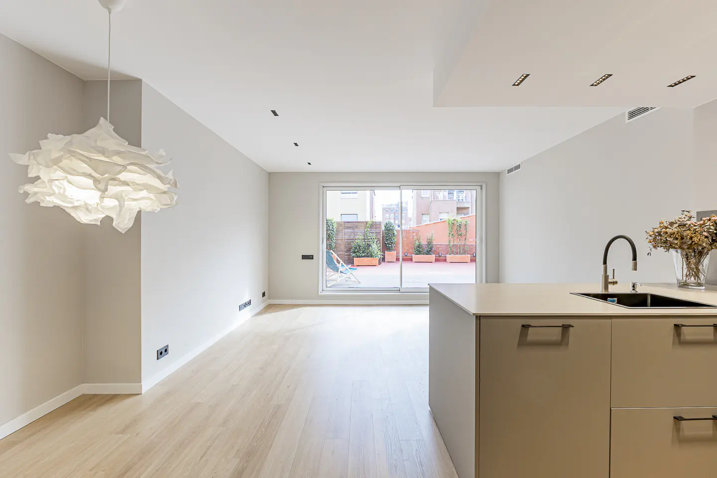 Bright, modern apartment interior with light wood floors, white walls, and a kitchen island. Sliding glass doors lead to a patio with plants.