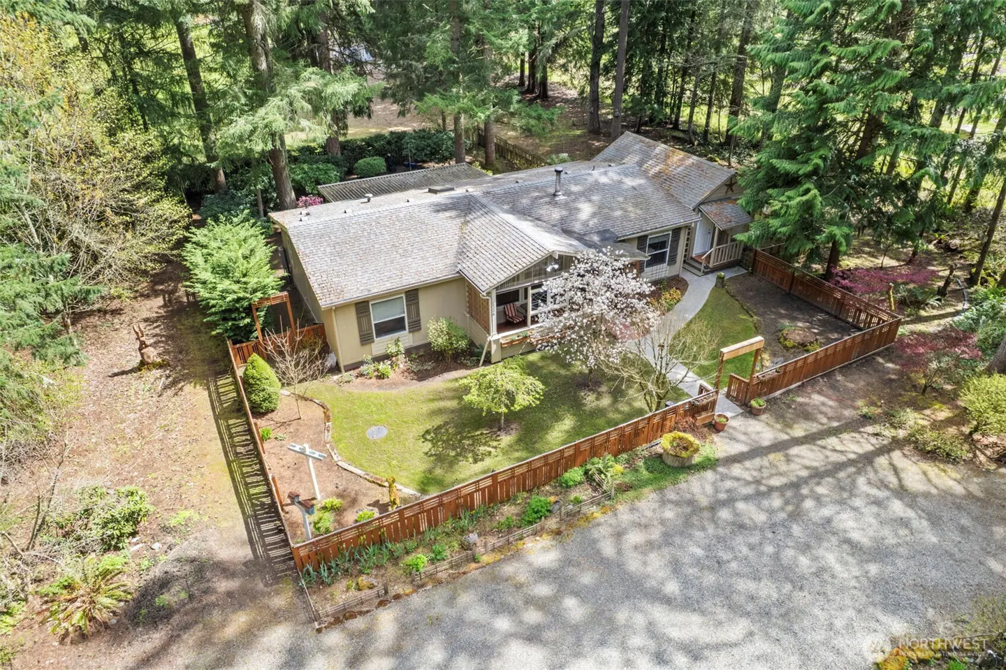 Aerial view of a beige house with a gray roof, surrounded by a wooden fence and tall green trees. A blooming tree stands in the front yard.