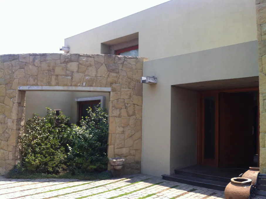 Modern home exterior with stone wall, arched opening with greenery, and a wooden door entrance. The house is painted in neutral tones.