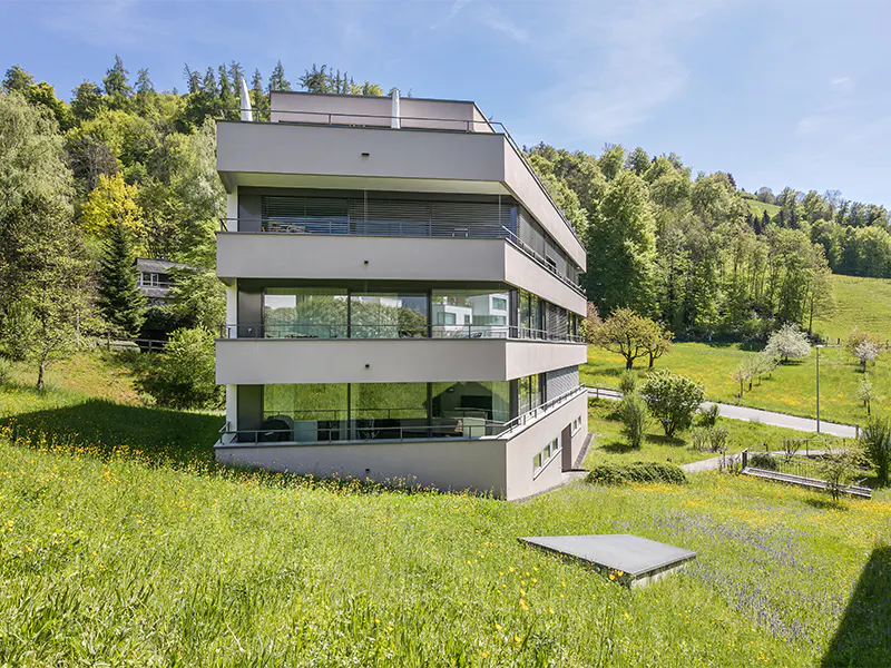 Modern three-story gray building with balconies, surrounded by a green meadow and trees under a blue sky.