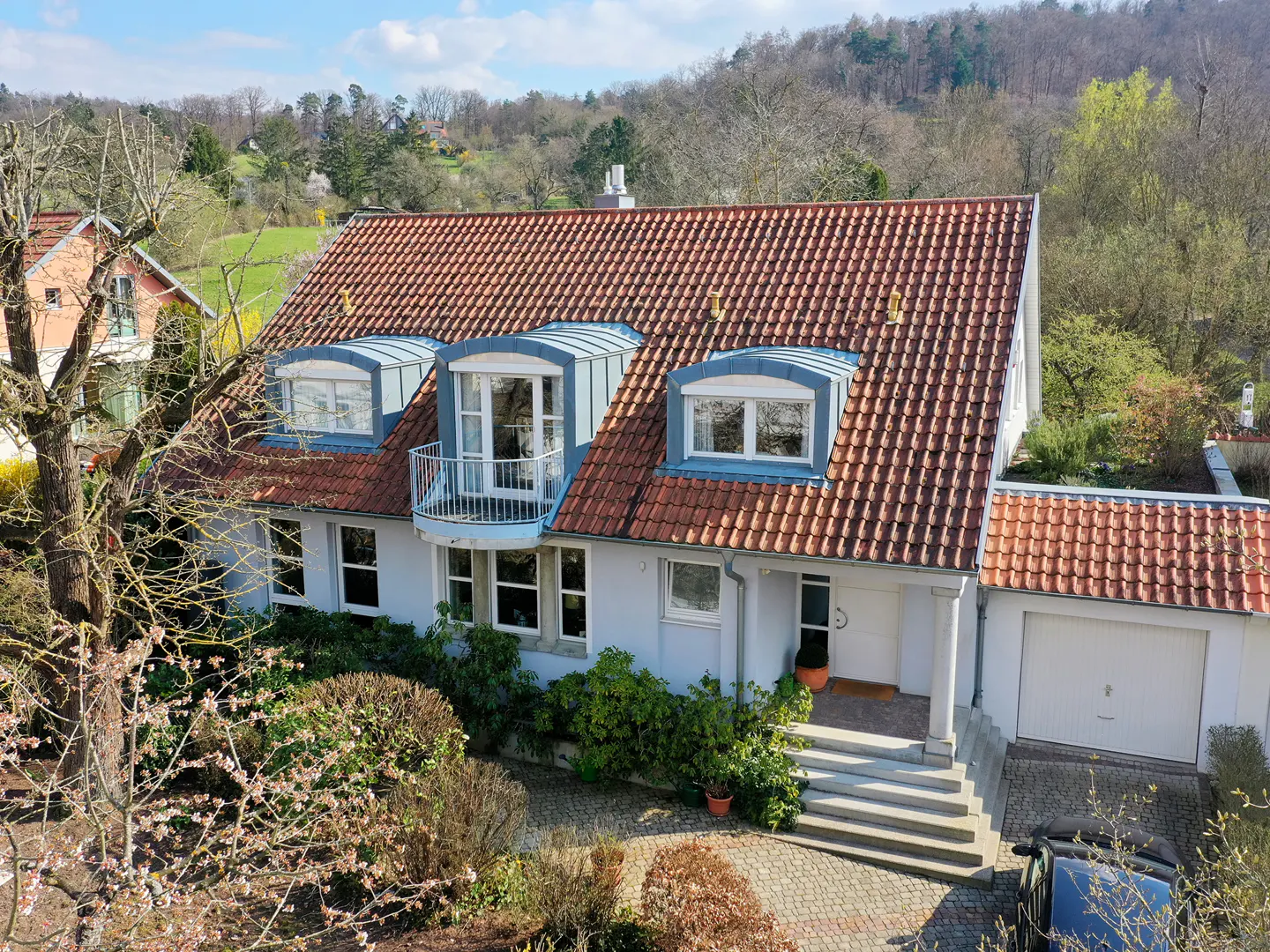 Aerial view of a two-story white house with a red tile roof, dormer windows, and a small balcony. Lush greenery surrounds the house.