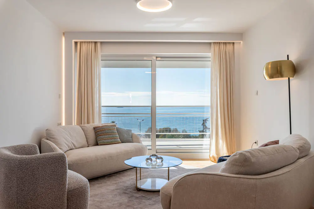 Bright living room with beige sofas, a glass table, and a view of the ocean through a large window with cream curtains. A gold lamp is on the right wall.