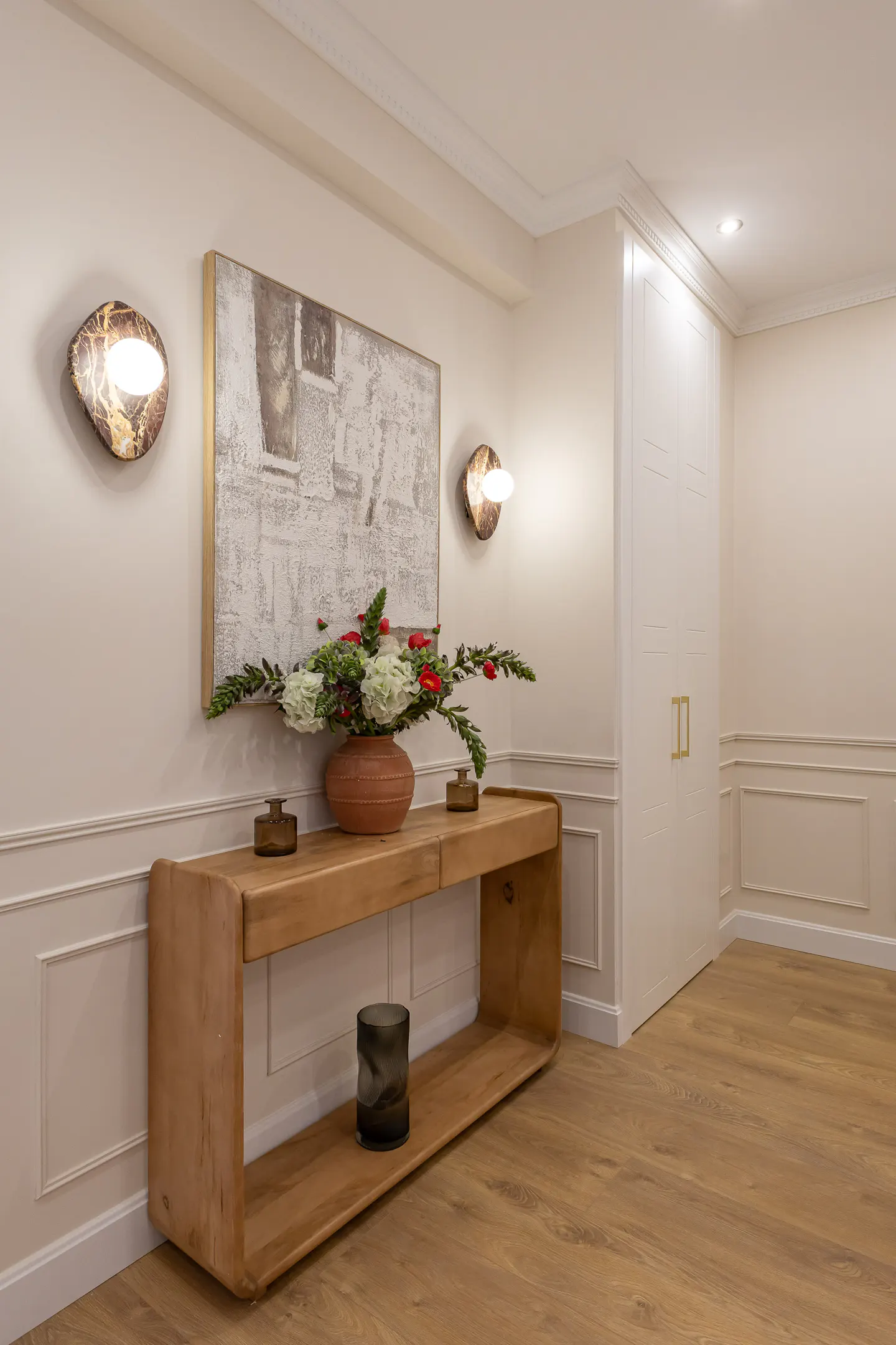 Entryway with a light wood console table, a floral arrangement, and abstract art. Two sconces flank the art. The walls are white with decorative molding. Wood floors.