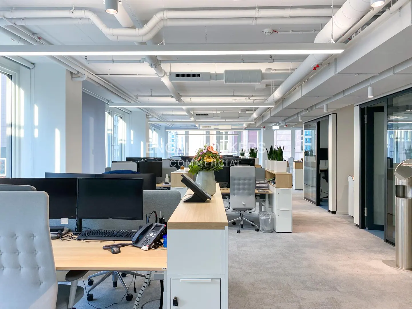 Bright, modern office space with desks, computers, and gray chairs. White ceiling with exposed pipes and linear lighting. Gray carpeted floor.