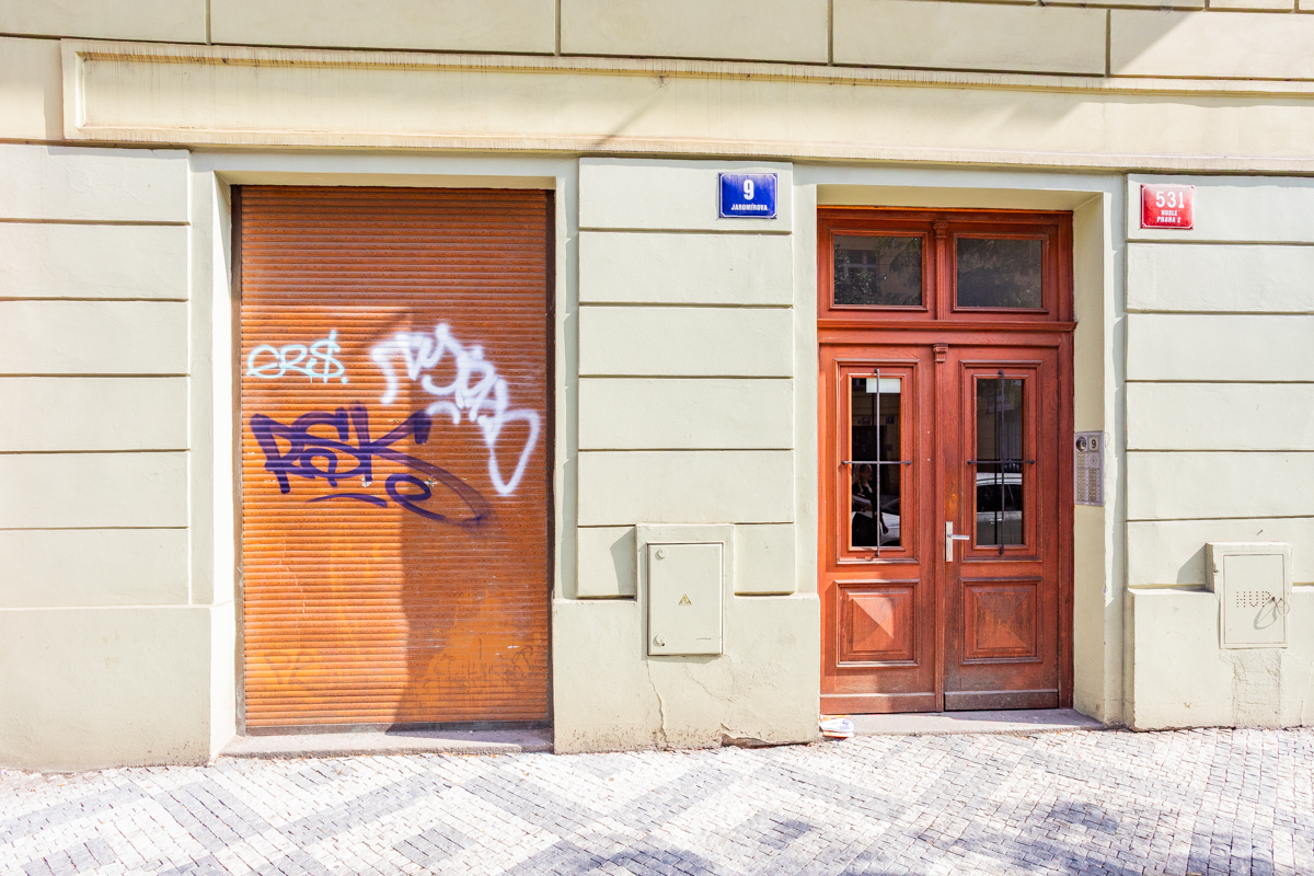 A beige building facade with a graffiti-covered orange metal shutter next to a classic wooden double door on a stone street.