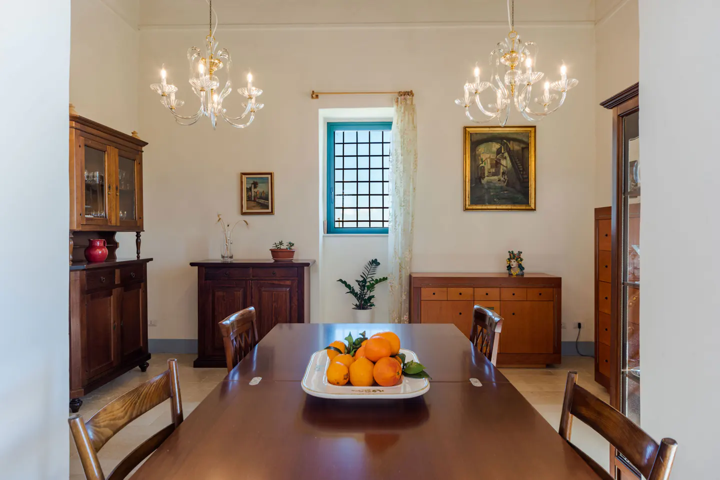 Dining room with a long wooden table, chairs, and a fruit platter. Two chandeliers hang from the ceiling. A window with a blue frame is in the background.