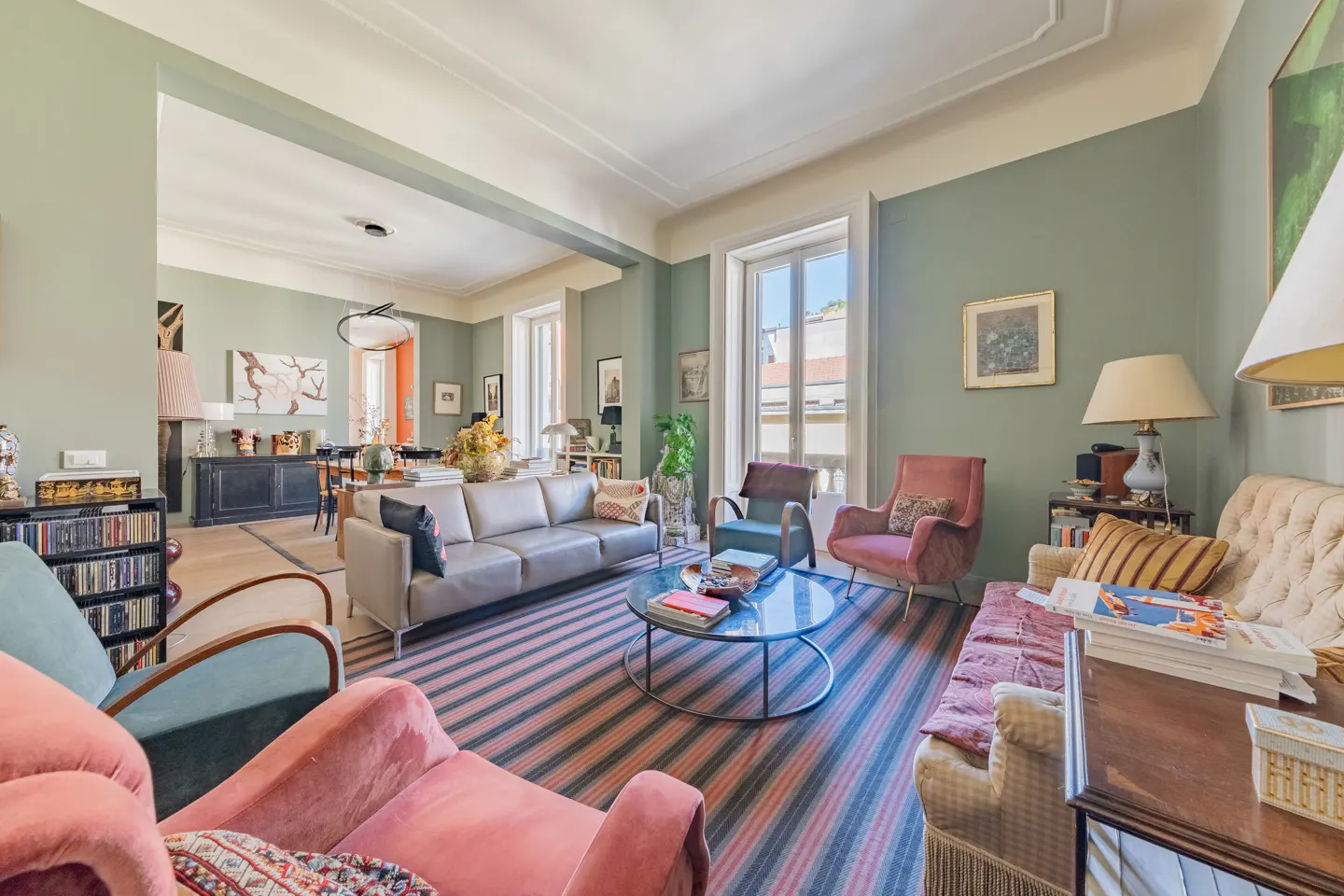 A spacious living room with sage green walls, striped rug, and an array of colorful furniture. Natural light streams through the windows.