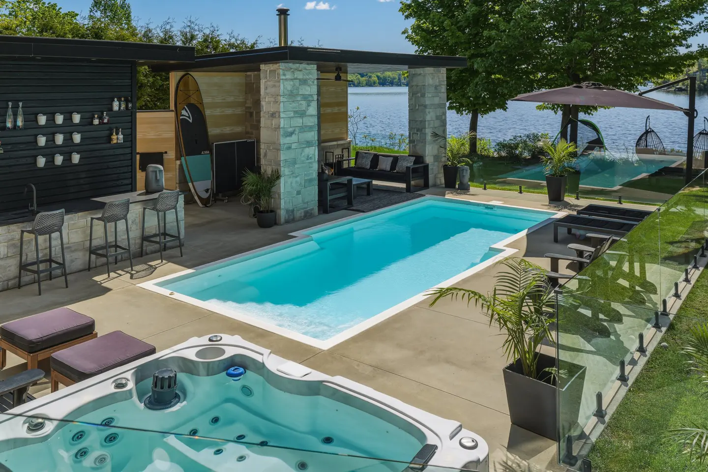 Luxury patio with a hot tub, swimming pool, outdoor bar, and lounge area overlooking a lake on a sunny day.