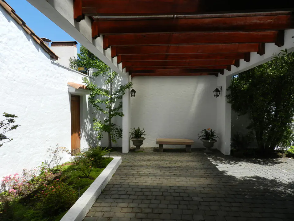 Covered walkway with a wooden pergola, white walls, and a stone path. A bench sits between potted plants. Landscaped garden on the left.