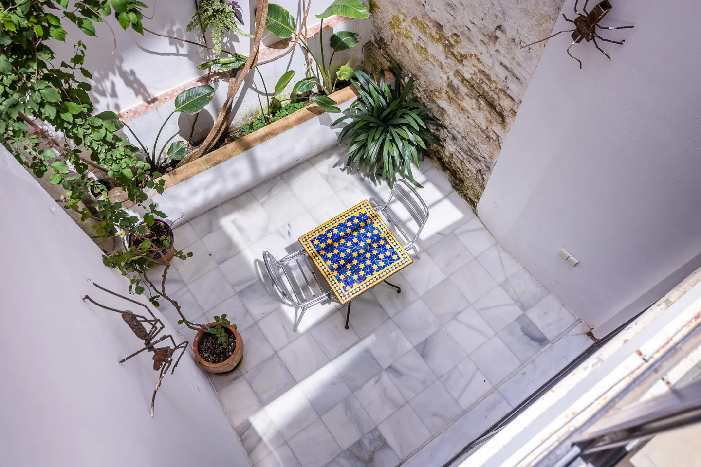 Overhead shot of a courtyard with a blue and yellow tiled table, two chairs, plants, and metal insect art.