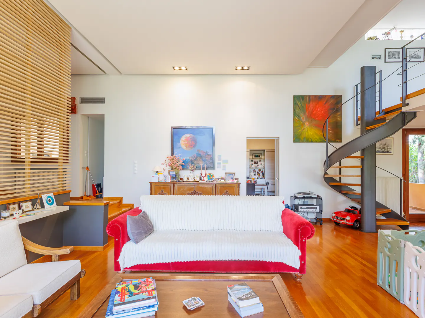 Bright living room with hardwood floors, a red sofa with a white cover, and a spiral staircase leading to the second floor.