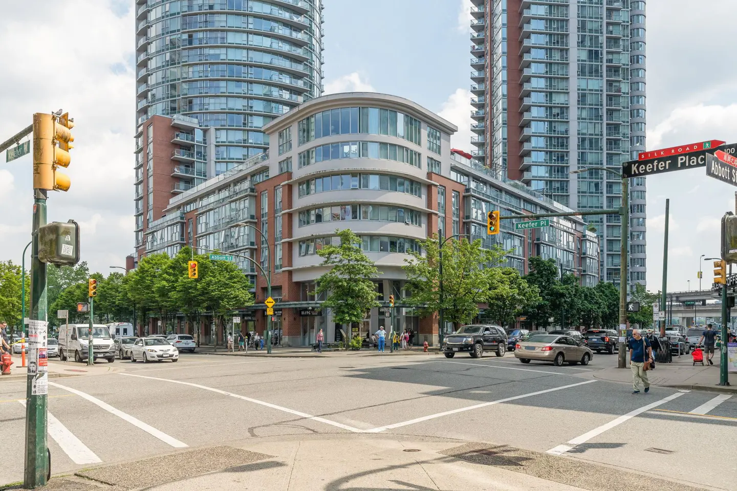 Street view of a city intersection with modern high-rise buildings. Cars and pedestrians are present. Traffic lights are visible.
