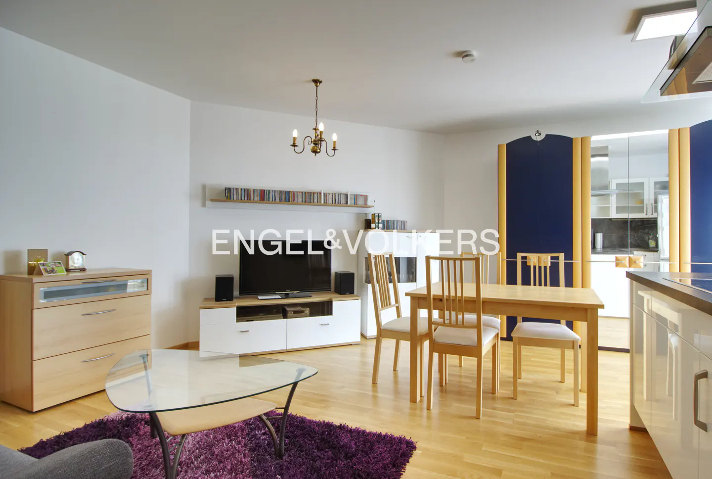 Living room with light wood floors, white walls, and a purple rug. A dining table with chairs sits near a TV and cabinet.