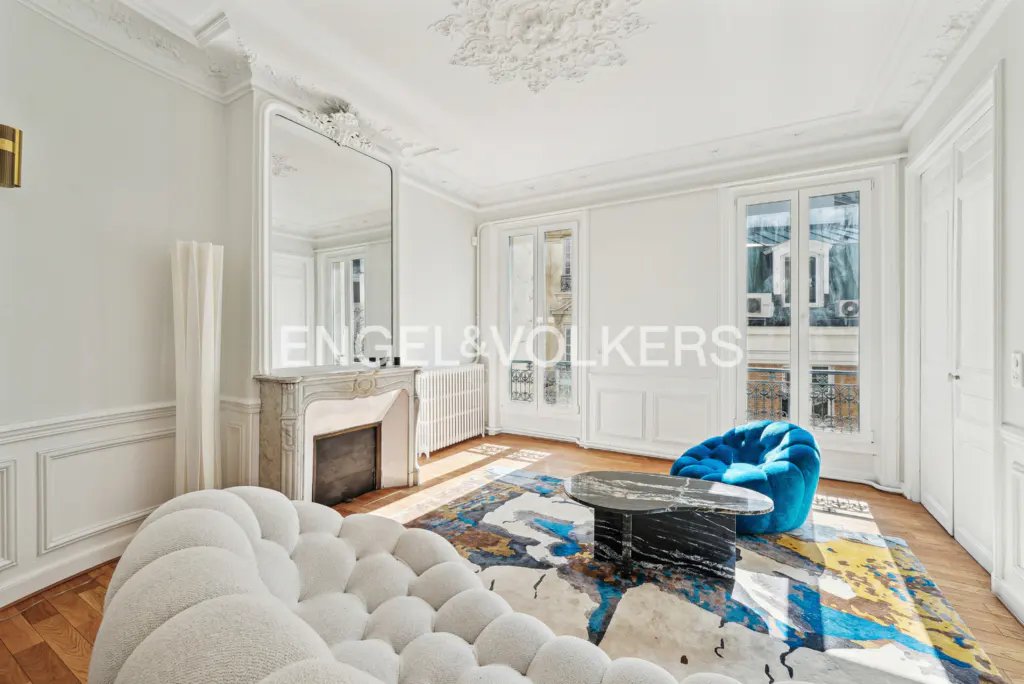 Bright living room with white walls, ornate ceiling, fireplace, and large mirror. A white bubble sofa and blue chair sit on a colorful rug.