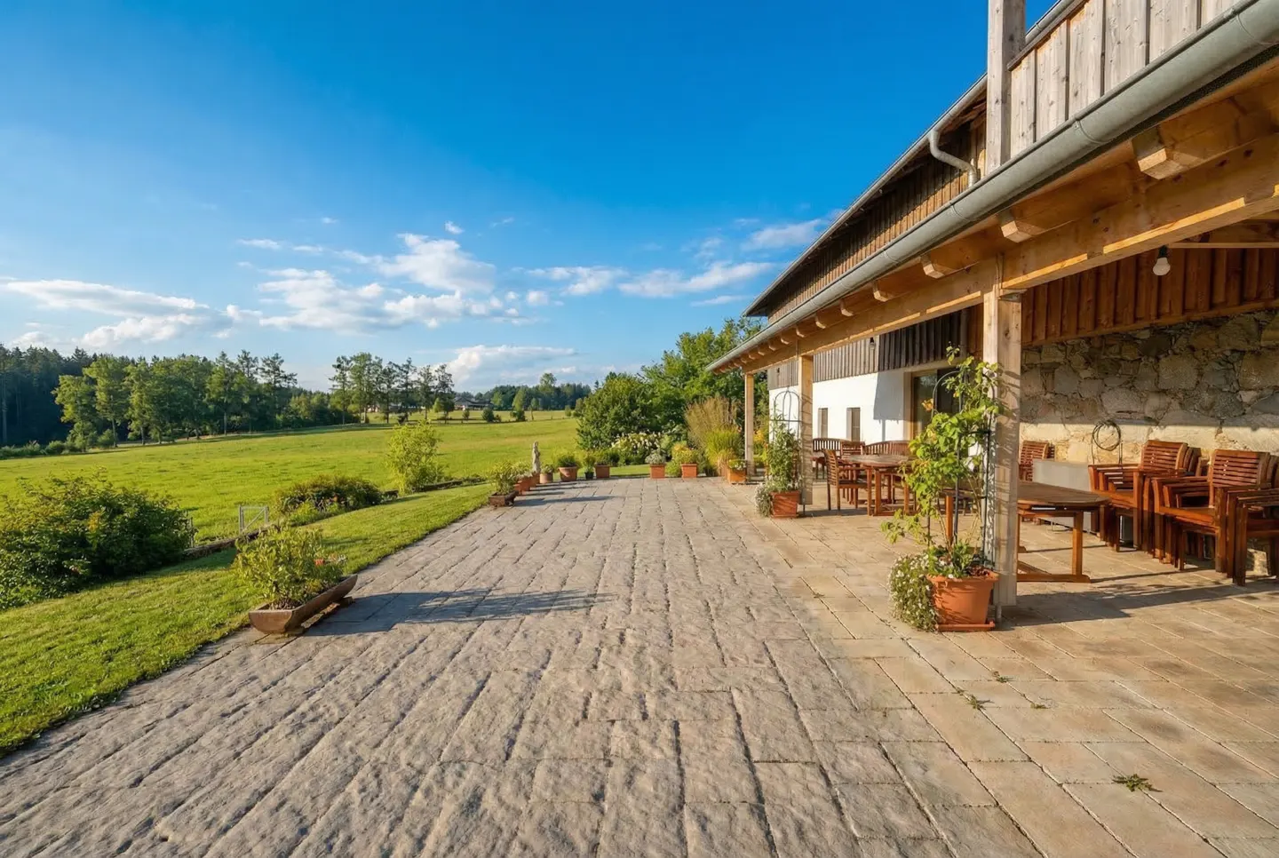 Stone patio with wooden tables and chairs outside a house, overlooking a green field under a blue sky.