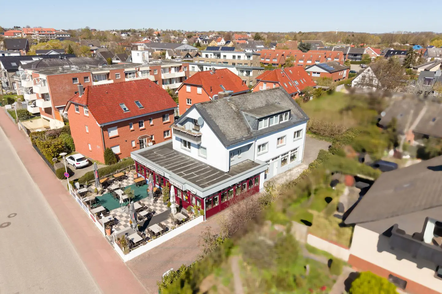 Aerial view of a white two-story building with a restaurant and outdoor seating area, surrounded by houses and trees.