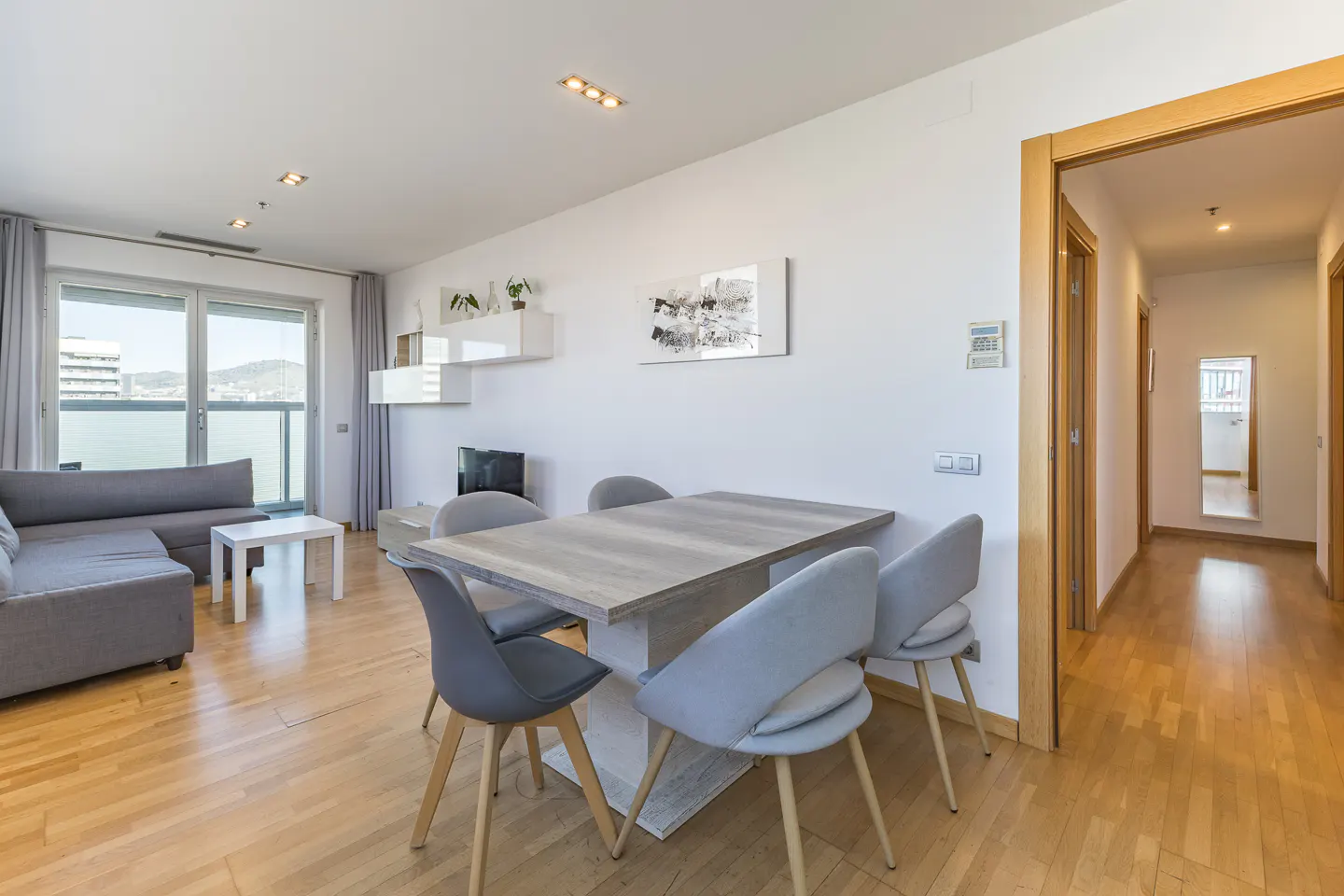 Bright apartment interior with wood floors, a gray sofa, a dining table with six chairs, and a hallway leading to other rooms.