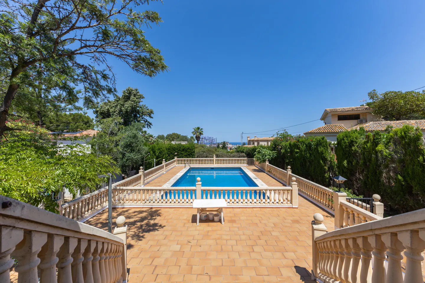 Outdoor pool with blue water, surrounded by a beige stone patio and railing, viewed from a staircase under a clear blue sky.