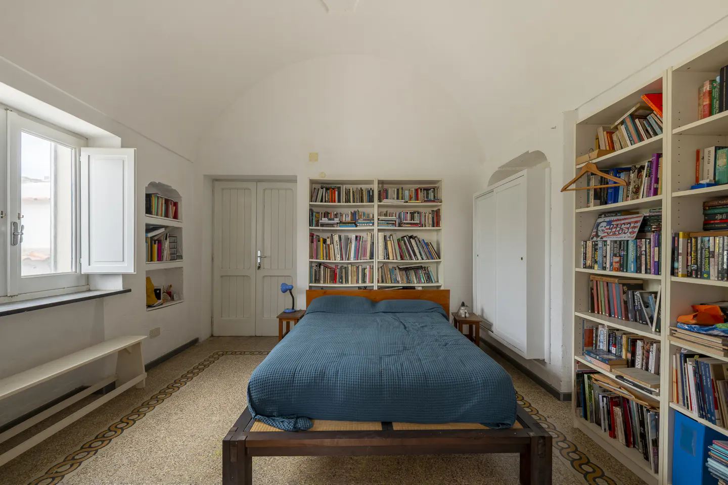 Bedroom with white walls, arched ceiling, and tiled floor. A bed with a blue coverlet is centered between bookshelves filled with books.