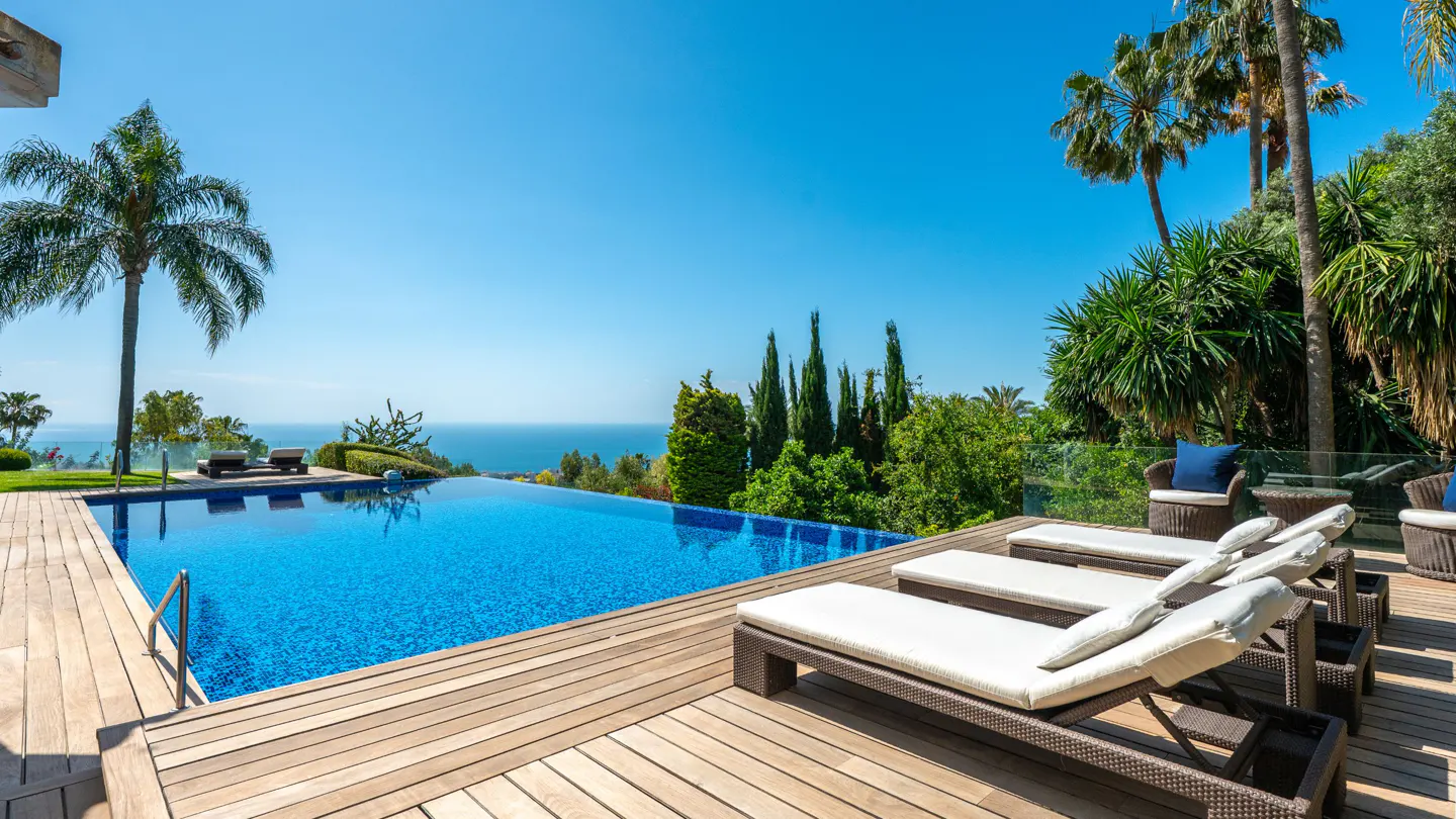 Infinity pool with blue tiles overlooking the ocean. Palm trees and lounge chairs on a wooden deck. Clear blue sky.