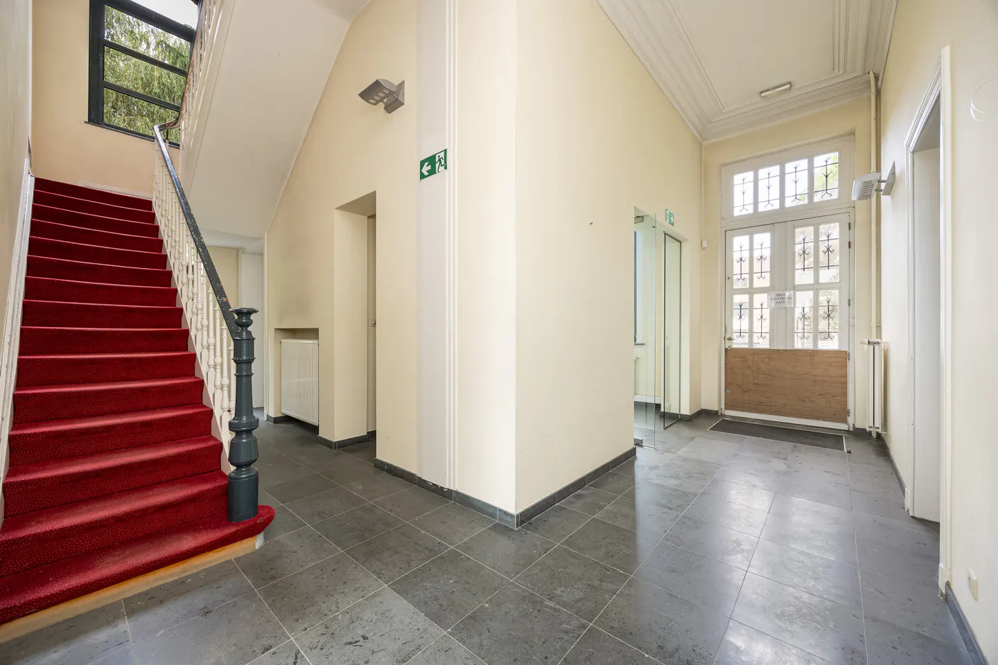 Bright building entrance with a red-carpeted staircase, grey tiled floor, and a glass-paned door with a wooden panel.