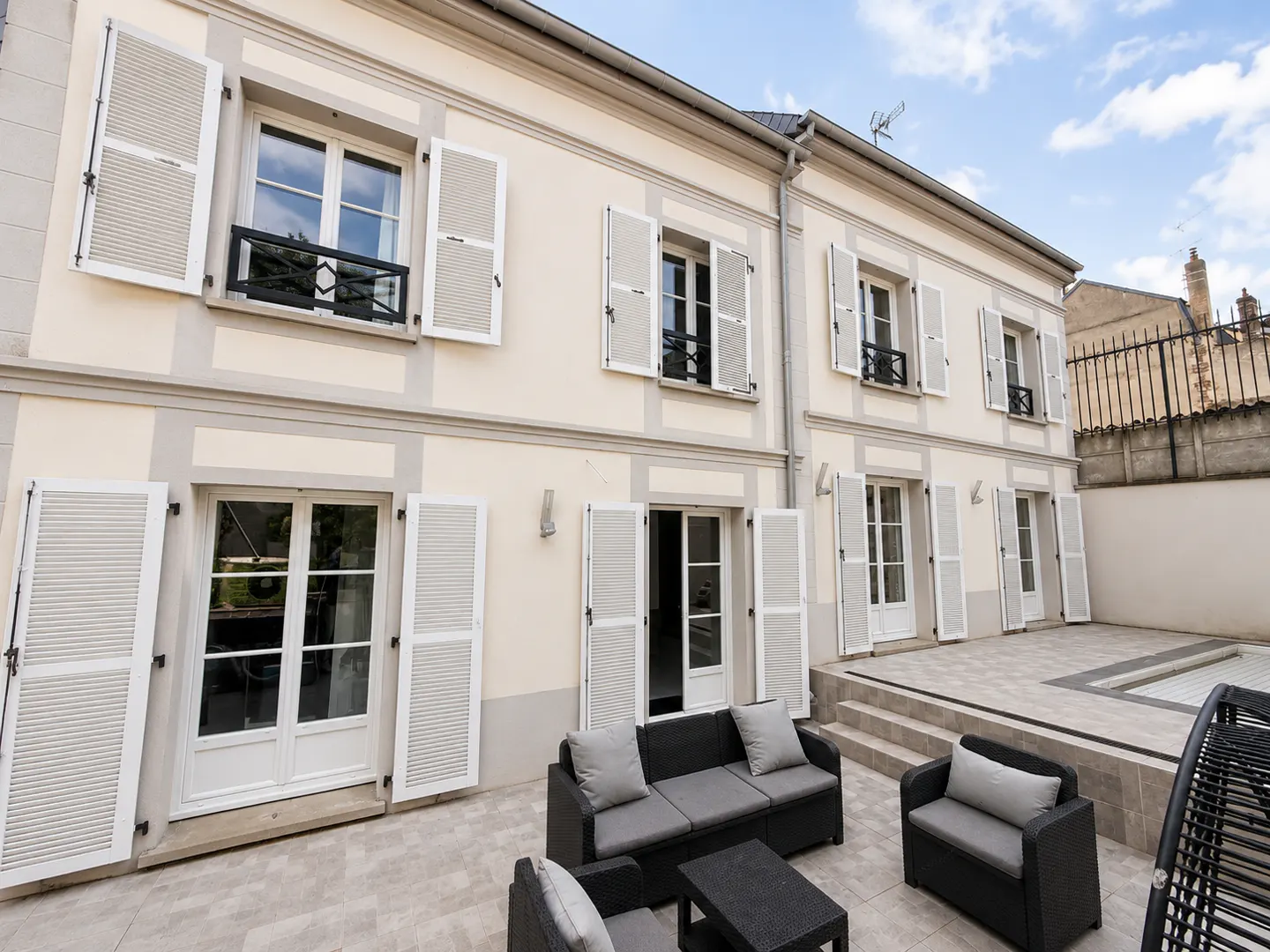 Cream-colored two-story house exterior with white shutters, glass doors, and a patio featuring dark wicker furniture.