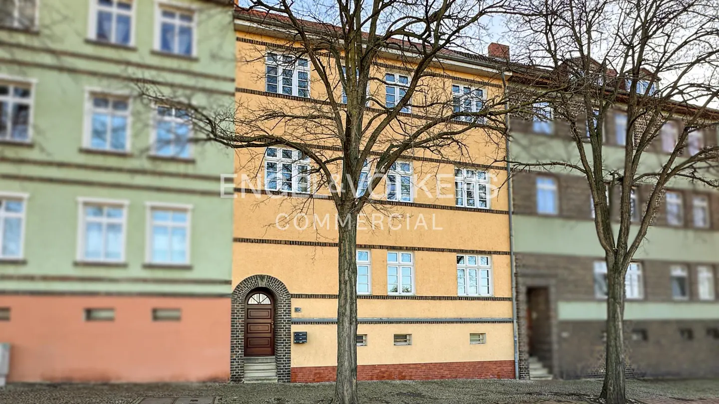 A three-story yellow building with white-framed windows, a brown door, and trees in front.