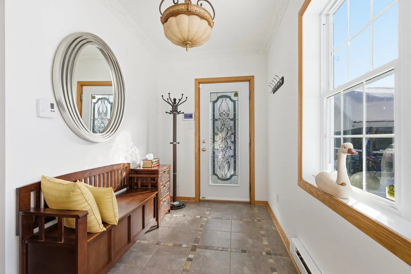 Bright foyer with white walls, tiled floor, and a wooden bench with yellow pillows. A decorative glass door and a window with a swan figurine are visible.