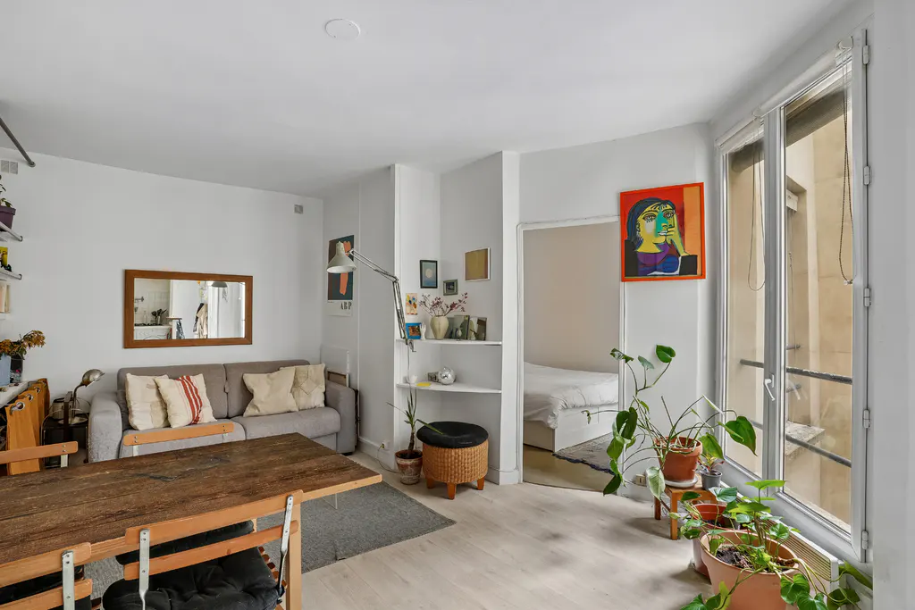 Bright apartment interior with a wooden table, gray sofa, and plants near a window. A colorful painting hangs on the wall.