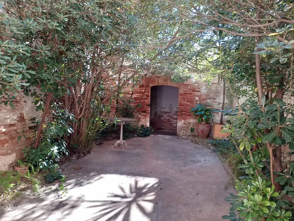 A secluded patio with a concrete floor, surrounded by lush trees, a small metal table, and an arched brick doorway.