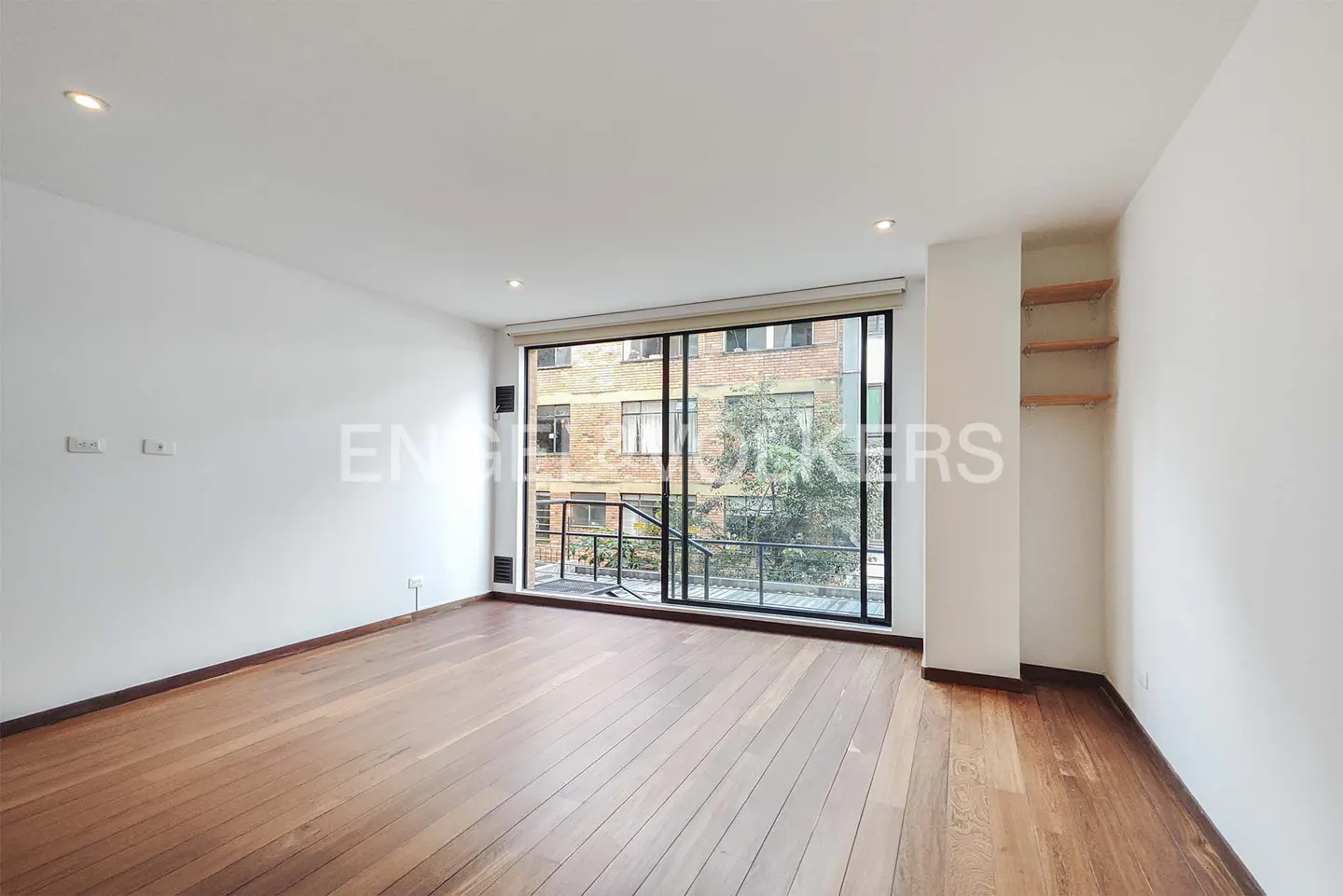 Empty room with white walls, hardwood floors, built-in wooden shelves, and a large window overlooking a brick building.