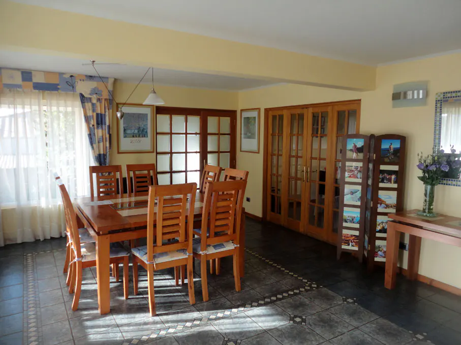 A dining room with a wooden table and chairs, tile floor, and yellow walls. Natural light streams in from a window with patterned curtains.