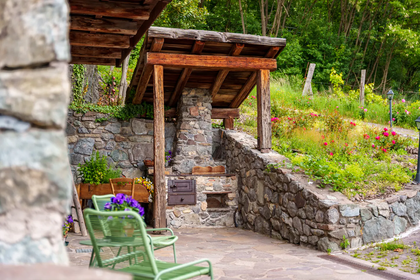 Stone patio with a wood-covered outdoor oven, green chairs, and a stone wall with flowers.