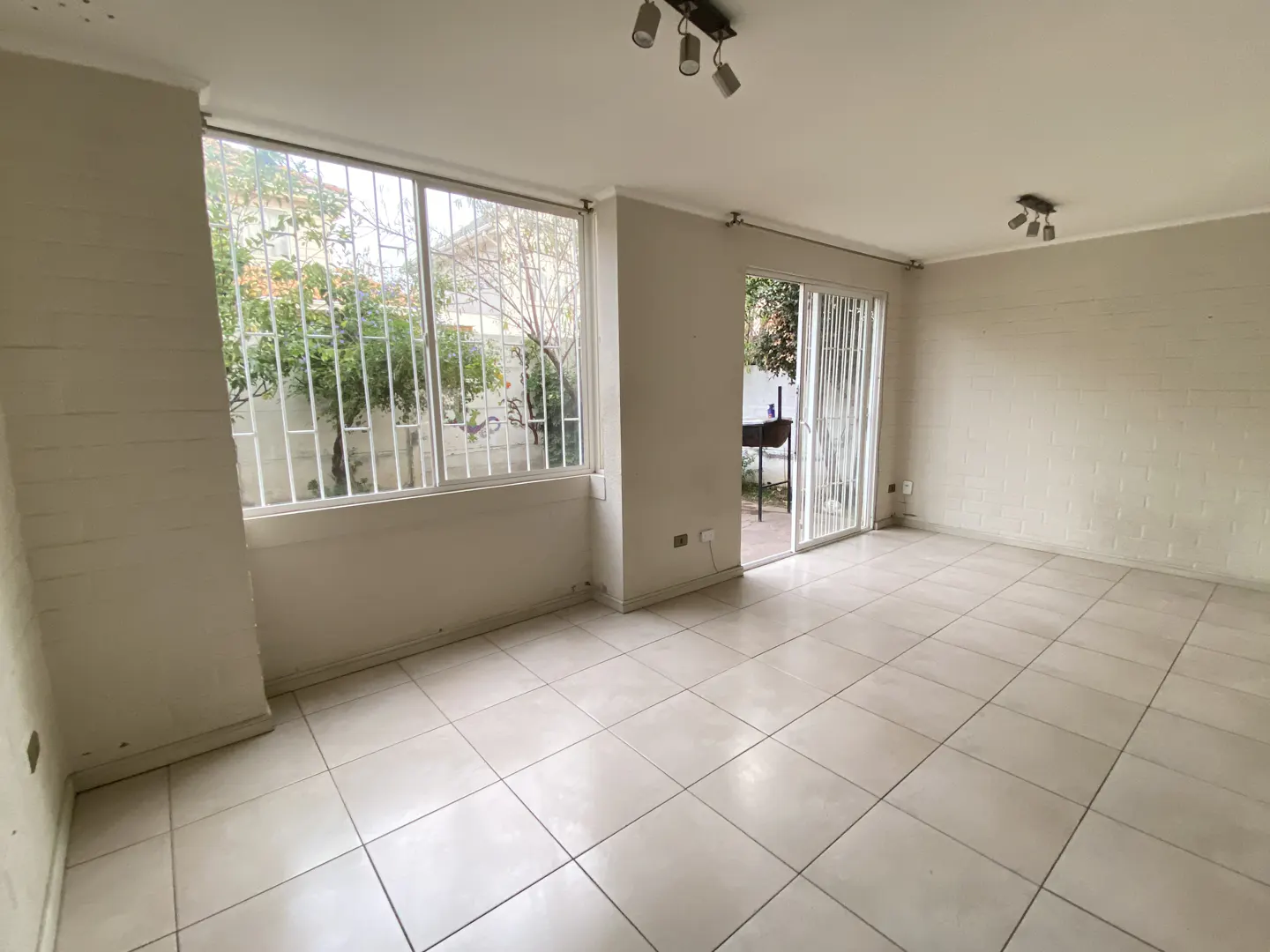 Bright, empty room with white tile floor, white walls, and a large window with a view of trees and a grill outside.