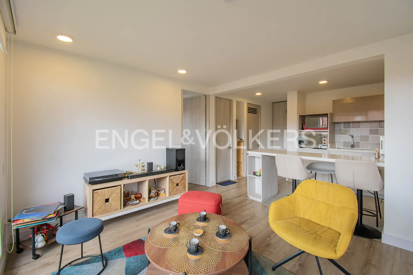 Modern living room with a yellow armchair, round coffee table, media console, and an open kitchen with a breakfast bar.