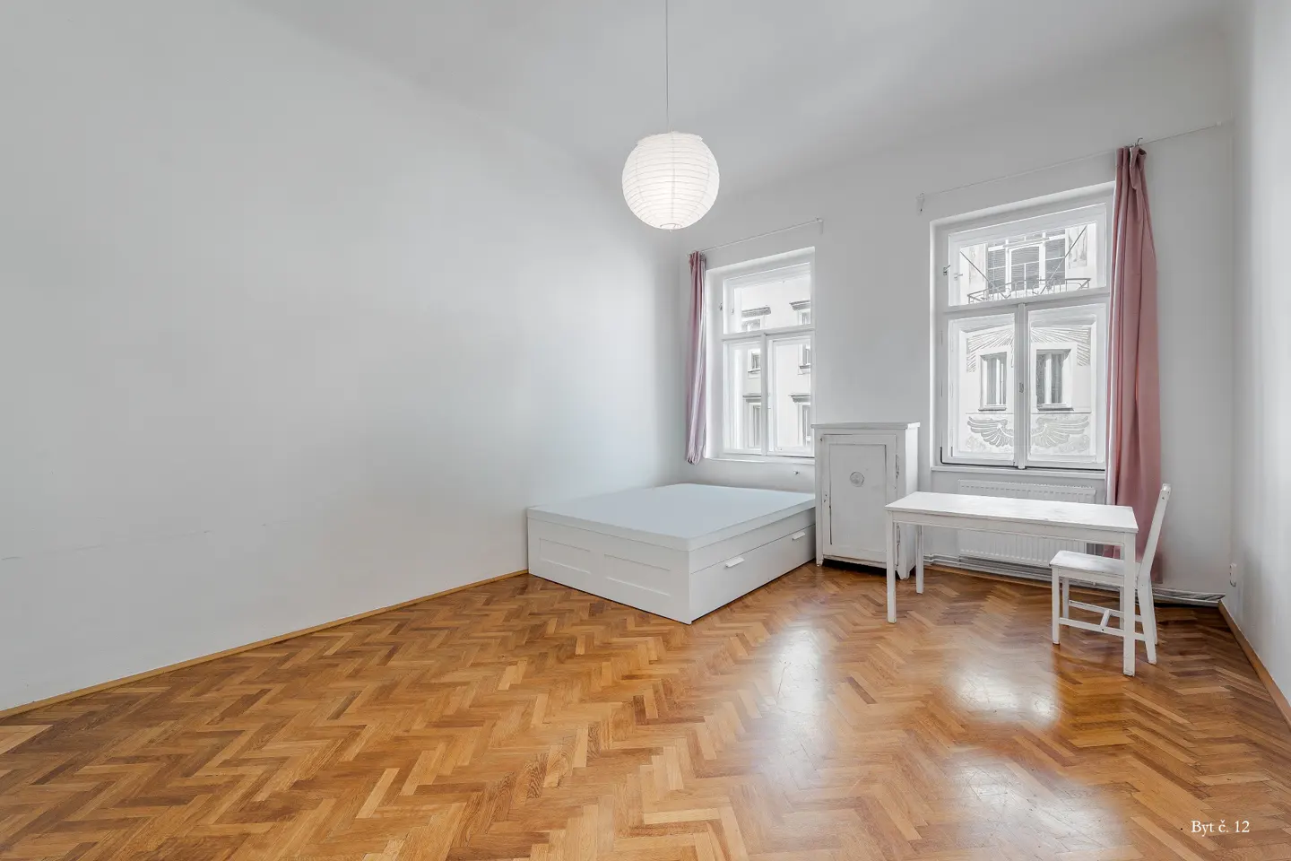 Bright room with herringbone wood floors, white bed, desk, chair, and pink curtains under a round paper pendant light.