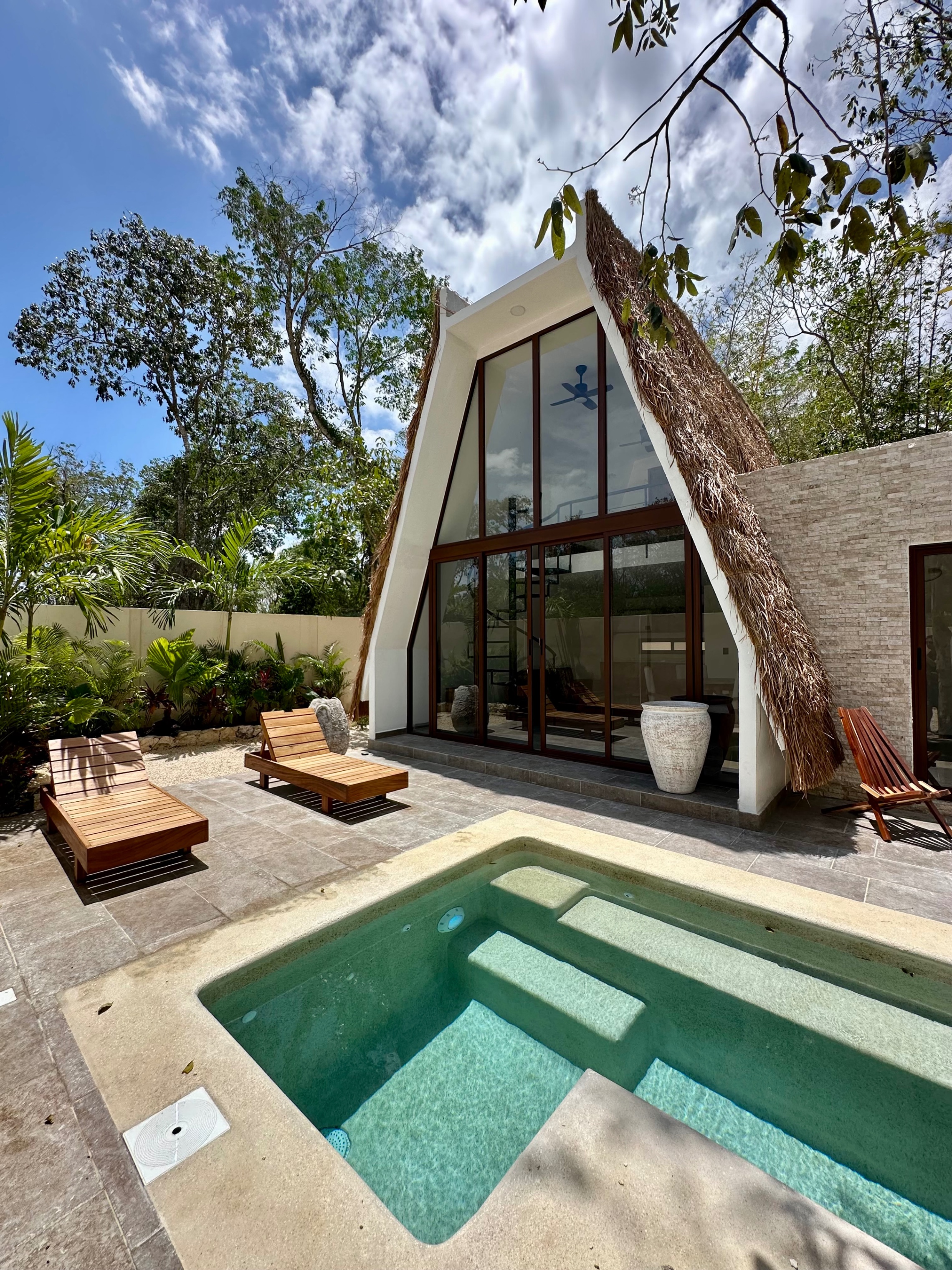 A-frame house with a thatched roof, a pool, and two wooden lounge chairs on a sunny day.