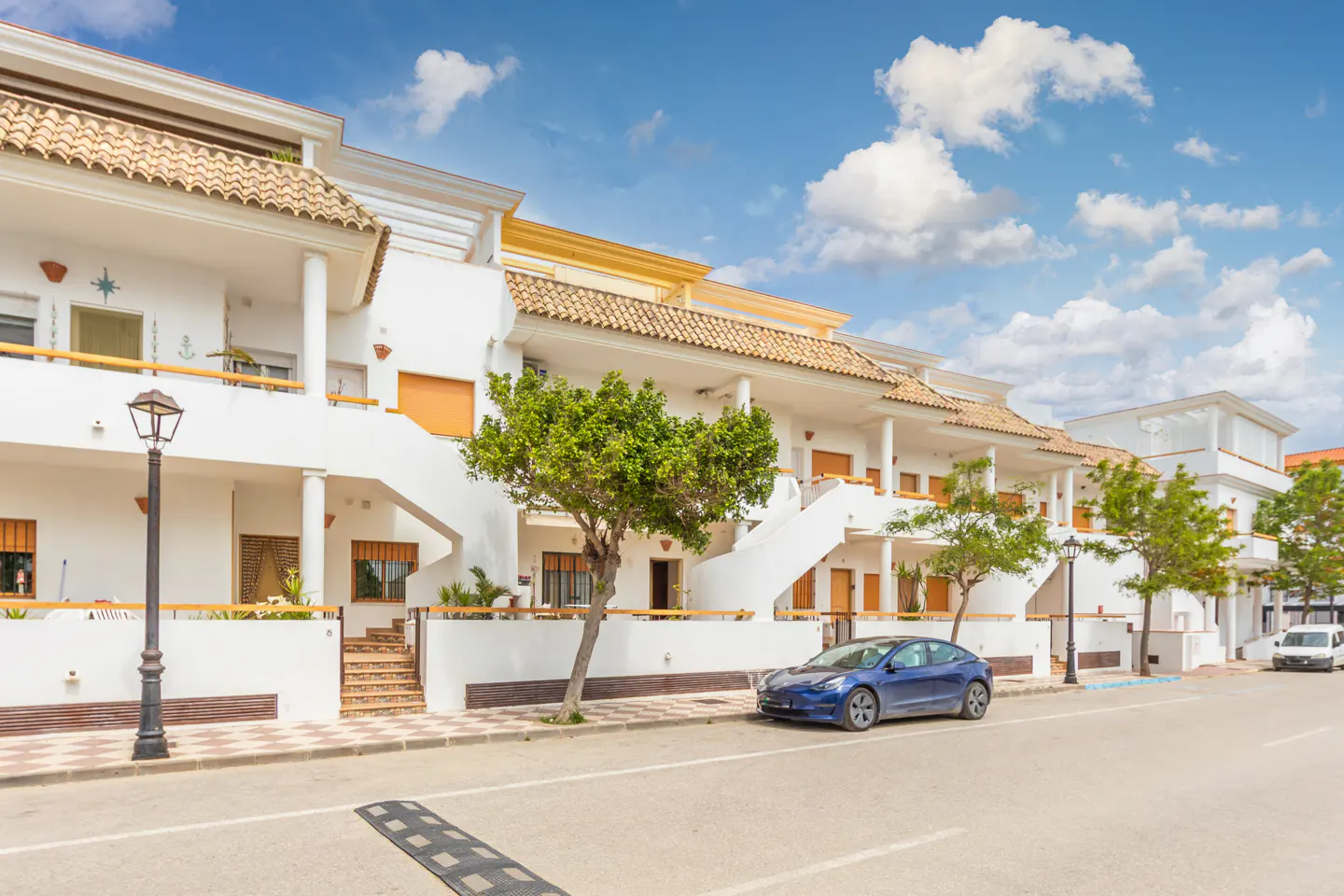 Row of white, multi-story townhouses with tile roofs under a blue, cloudy sky. A blue car is parked on the street.