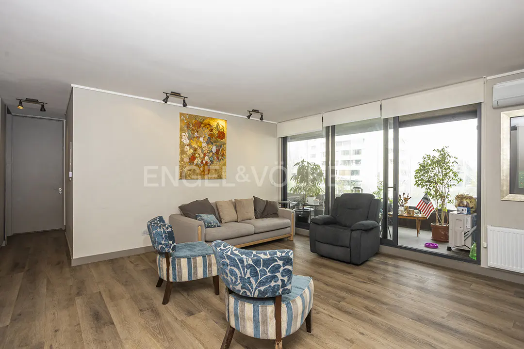 A living room with a gray sofa, blue chairs, and a balcony with plants and an American flag.