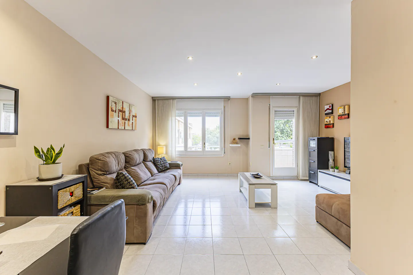 A living room with a brown sofa, white tile floor, and beige walls. A window and door let in natural light.