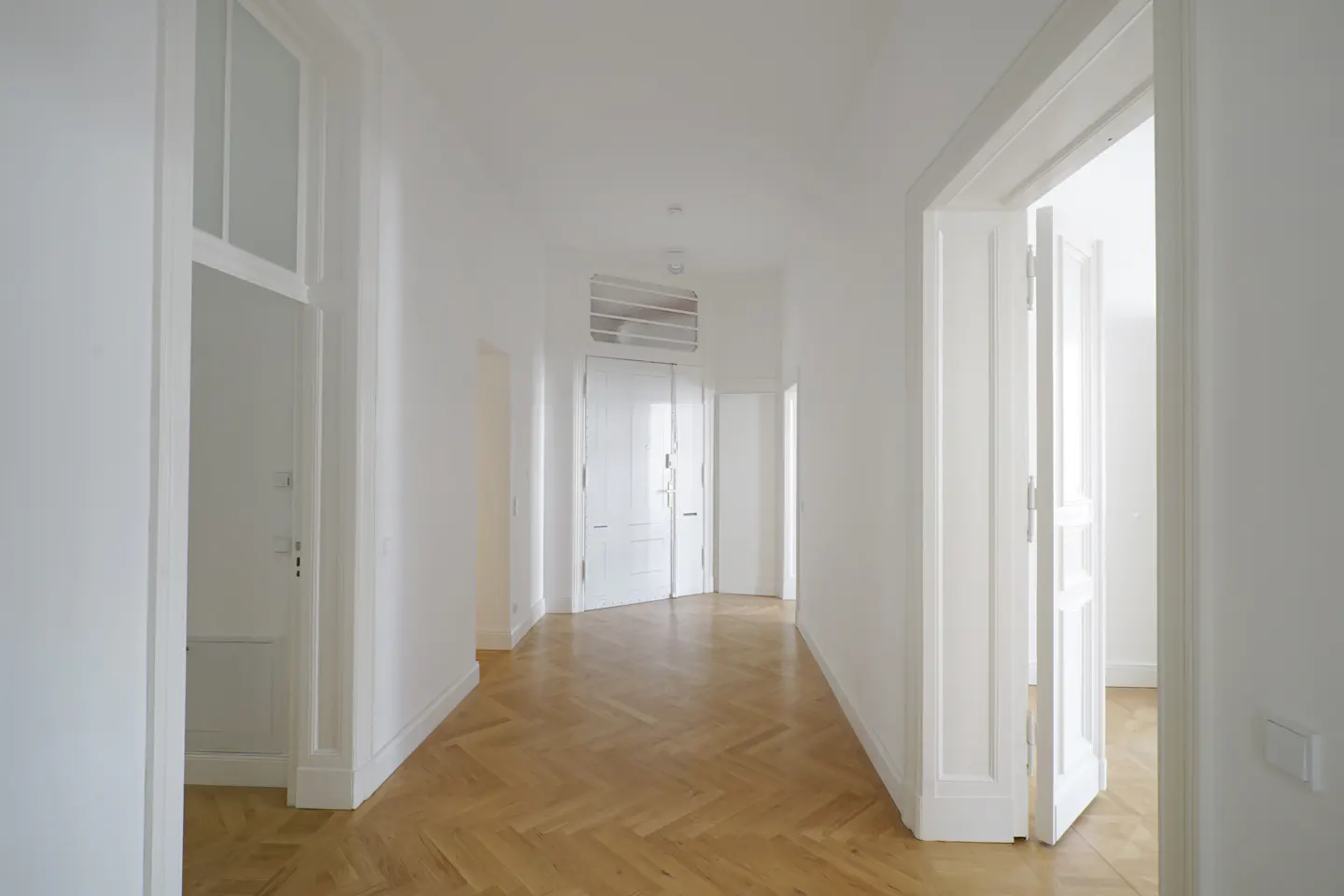 Bright, white hallway with herringbone wood floors. White doors line the hall, some open, leading to other rooms.
