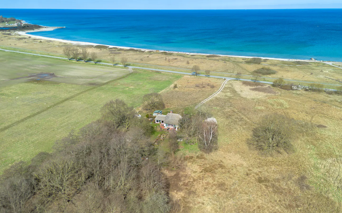 Aerial view of a cottage with a thatched roof near a beach and blue ocean. Fields and trees surround the house.